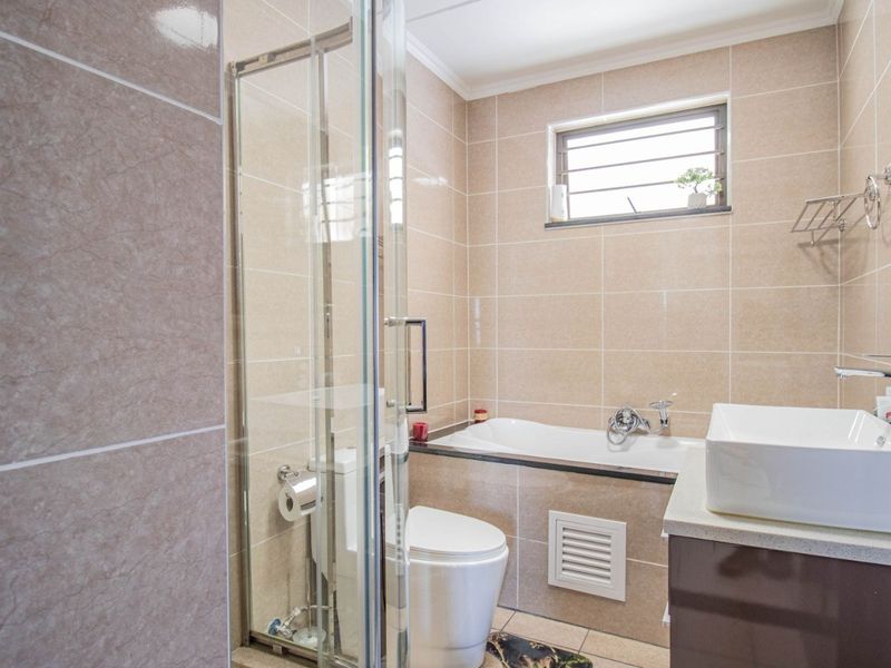 Modern bathroom featuring a glass shower enclosure, bathtub, vanity basin, and neutral floor-to-ceiling tiling with a louvered window for natural ventilation.