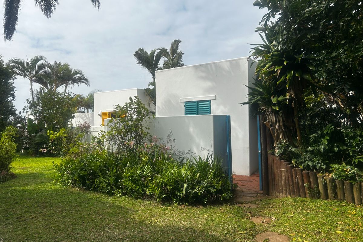 A view of the rear exterior of the apartment, featuring white rendered walls, colorful shutters, and a landscaped garden area with a paved path.