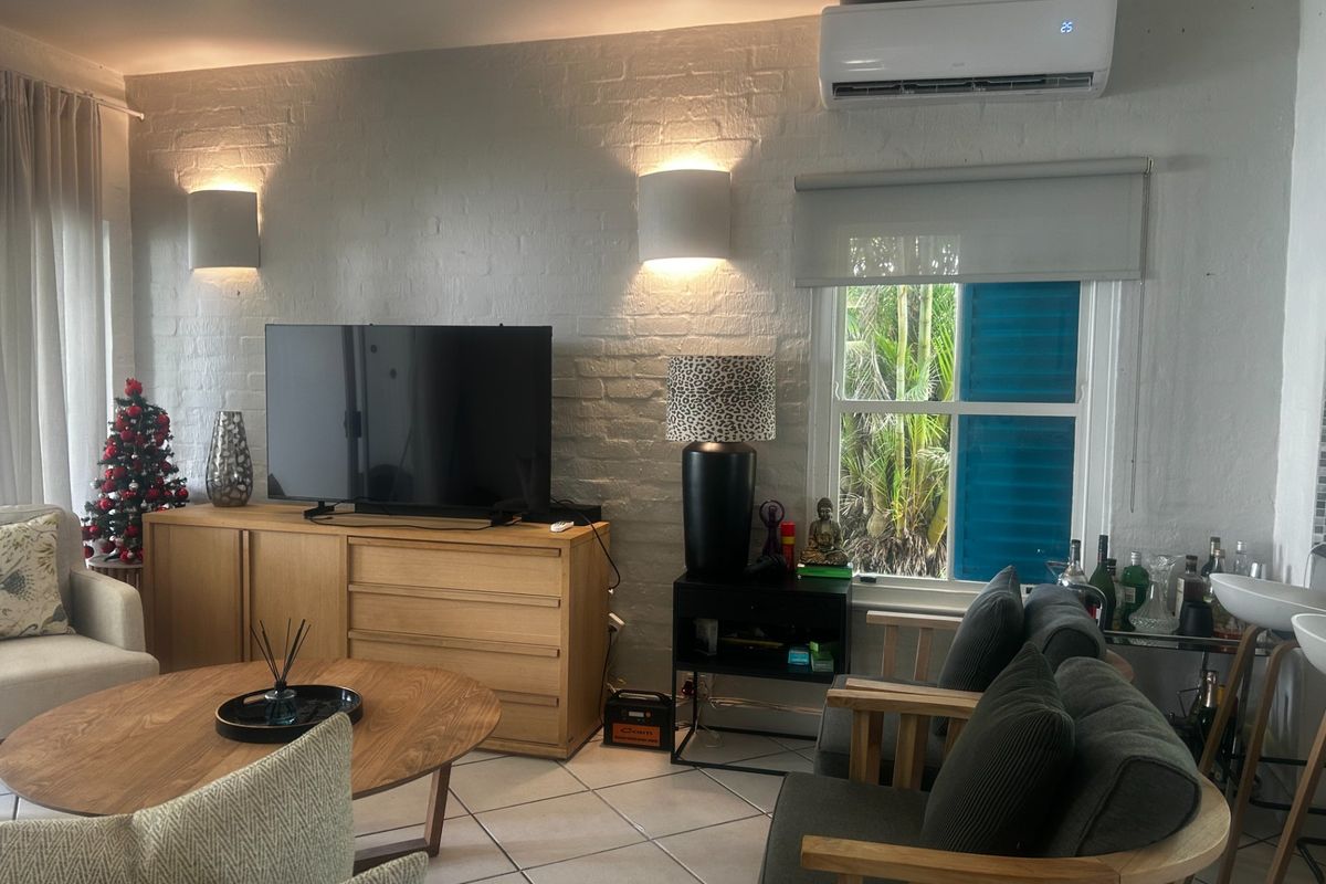 A comfortable living room featuring a painted brick feature wall, split-system air conditioning, tiled flooring, and natural light through a sash window.