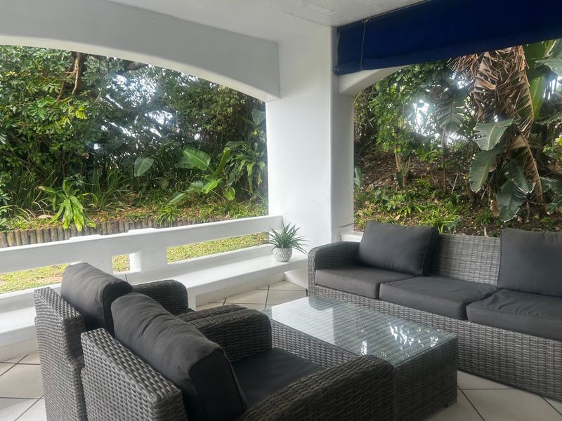 A covered patio area featuring tiled flooring, a built-in concrete balustrade, and a retractable shade blind, overlooking a lush garden setting.