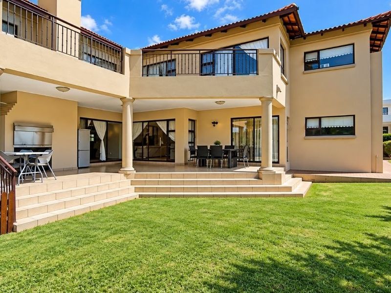 Spacious rear exterior featuring a large tiled patio with a built-in braai, upper-level balcony, and well-maintained garden area.