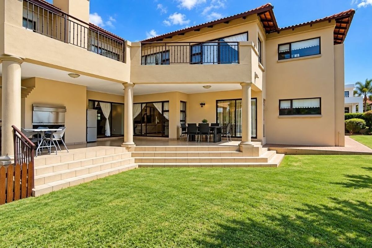 Spacious rear exterior featuring a large tiled patio with a built-in braai, upper-level balcony, and well-maintained garden area.