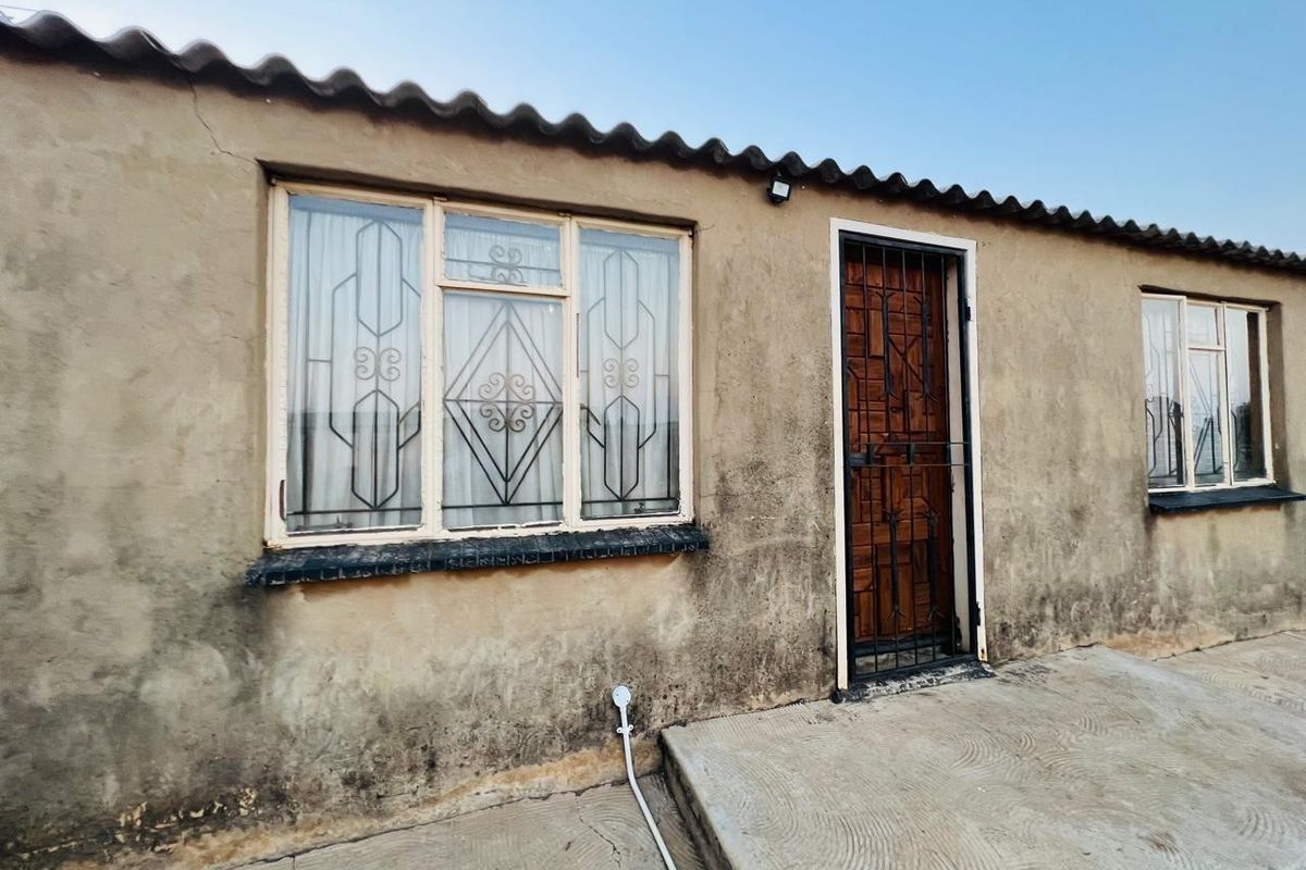 Exterior view of the rear facade featuring weathered plaster walls, security gates, and metal window frames with a concrete patio area.