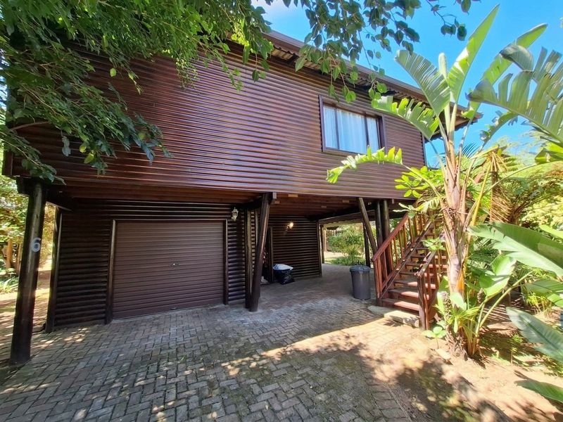 Front elevation of a stilted timber-clad apartment building featuring a ground-level garage, paved driveway, and an external staircase leading to the upper level.