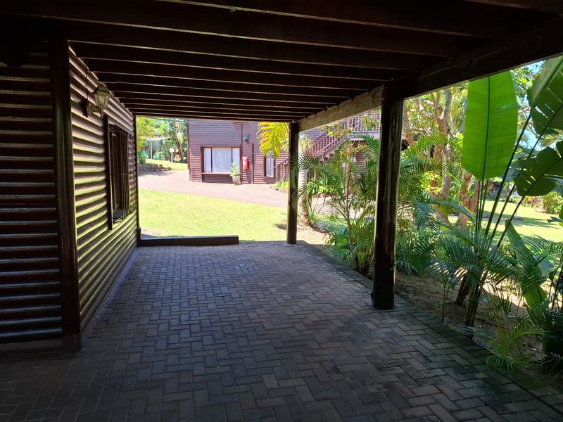A covered outdoor patio area featuring timber beam ceilings, support posts, and brick paving, overlooking a landscaped garden setting.