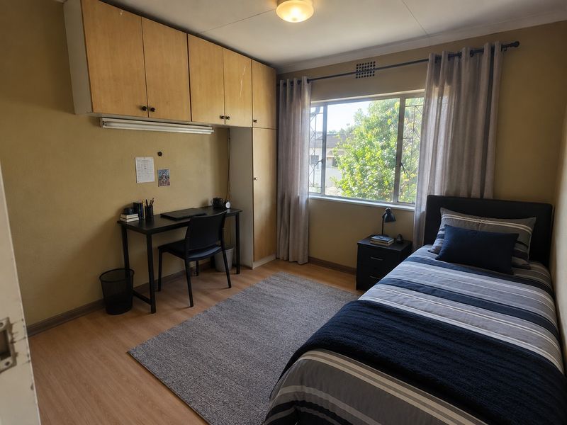 A functional bedroom featuring built-in wooden cabinetry, laminate flooring, and a window with security bars, offering a practical layout for a study and sleeping area.