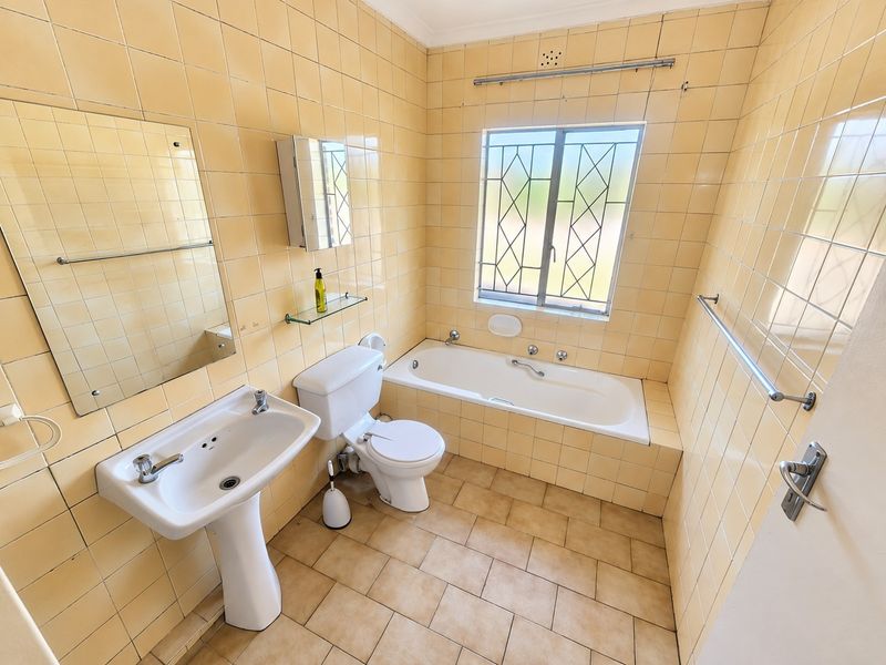 A functional bathroom featuring a pedestal basin, toilet, and built-in bathtub with full-height wall tiling and a security-fitted window.