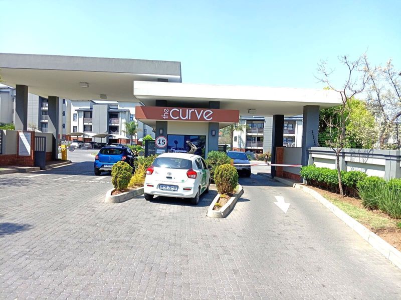 The entrance to The Curve apartment complex, featuring a paved driveway, security boom, and modern concrete canopy structure.