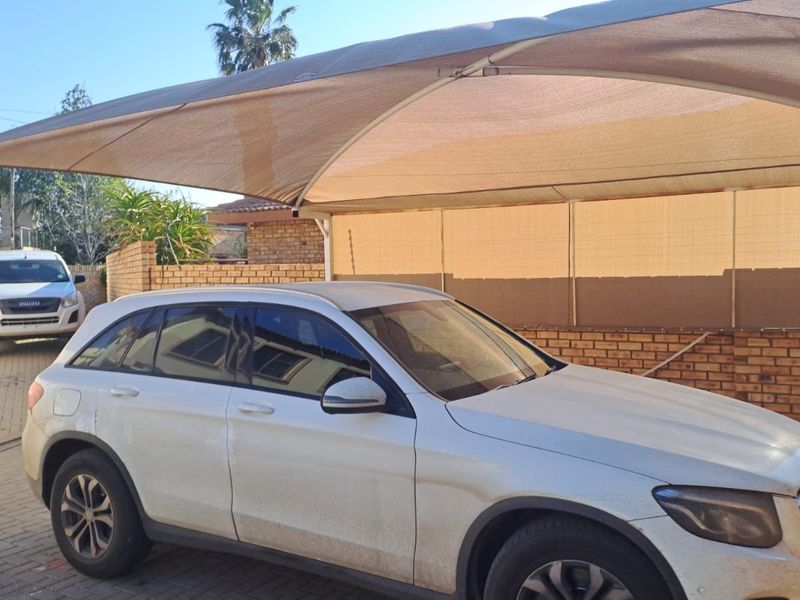 Front exterior area featuring a paved driveway, brick boundary wall with electric fencing, and a large shade sail structure for vehicle protection.