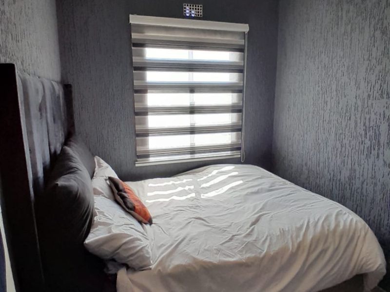 A compact bedroom featuring dark textured wallpaper, tiled flooring, and a window fitted with modern roller blinds.