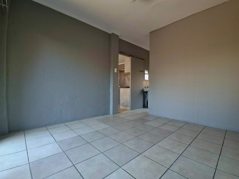 An empty living area featuring neutral grey walls and ceramic floor tiles, with a view through to an adjacent kitchen space.