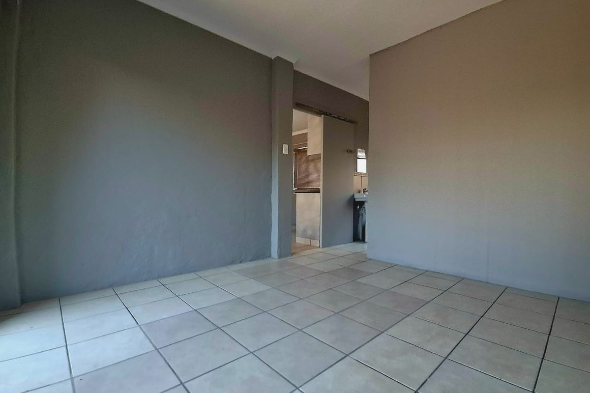An empty living area featuring neutral grey walls and ceramic floor tiles, with a view through to an adjacent kitchen space.