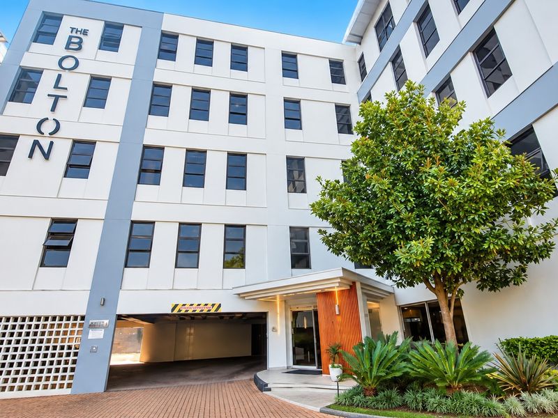 Modern apartment building facade featuring a paved driveway, secure undercover parking entrance, and well-maintained landscaped garden area.