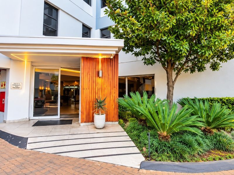 Modern apartment building entrance featuring a glass door, timber slat feature wall, recessed lighting, and well-maintained landscaped garden area.