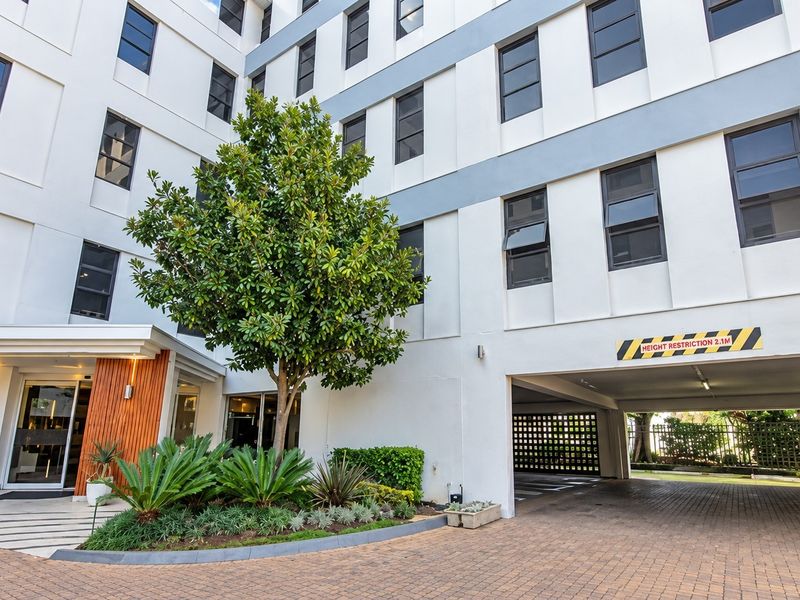 Front exterior view of the apartment building featuring a paved driveway, undercover parking area, and landscaped garden beds.