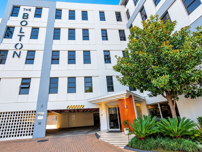 Front exterior view of The Bolton apartment complex, featuring a paved driveway, undercover parking entrance, and well-maintained landscaped garden area.