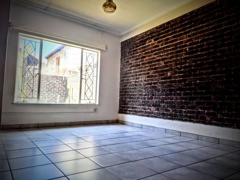 An empty living room featuring a prominent exposed brick feature wall, ceramic tiled flooring, and large windows with security burglar bars.