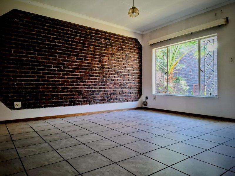 An empty living room featuring a prominent exposed brick wall, ceramic tiled flooring, and a large window with security bars.