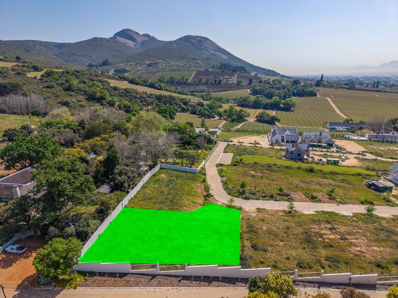 Aerial view of a vacant residential plot featuring a boundary wall and access to a paved road, set against a scenic mountain backdrop.