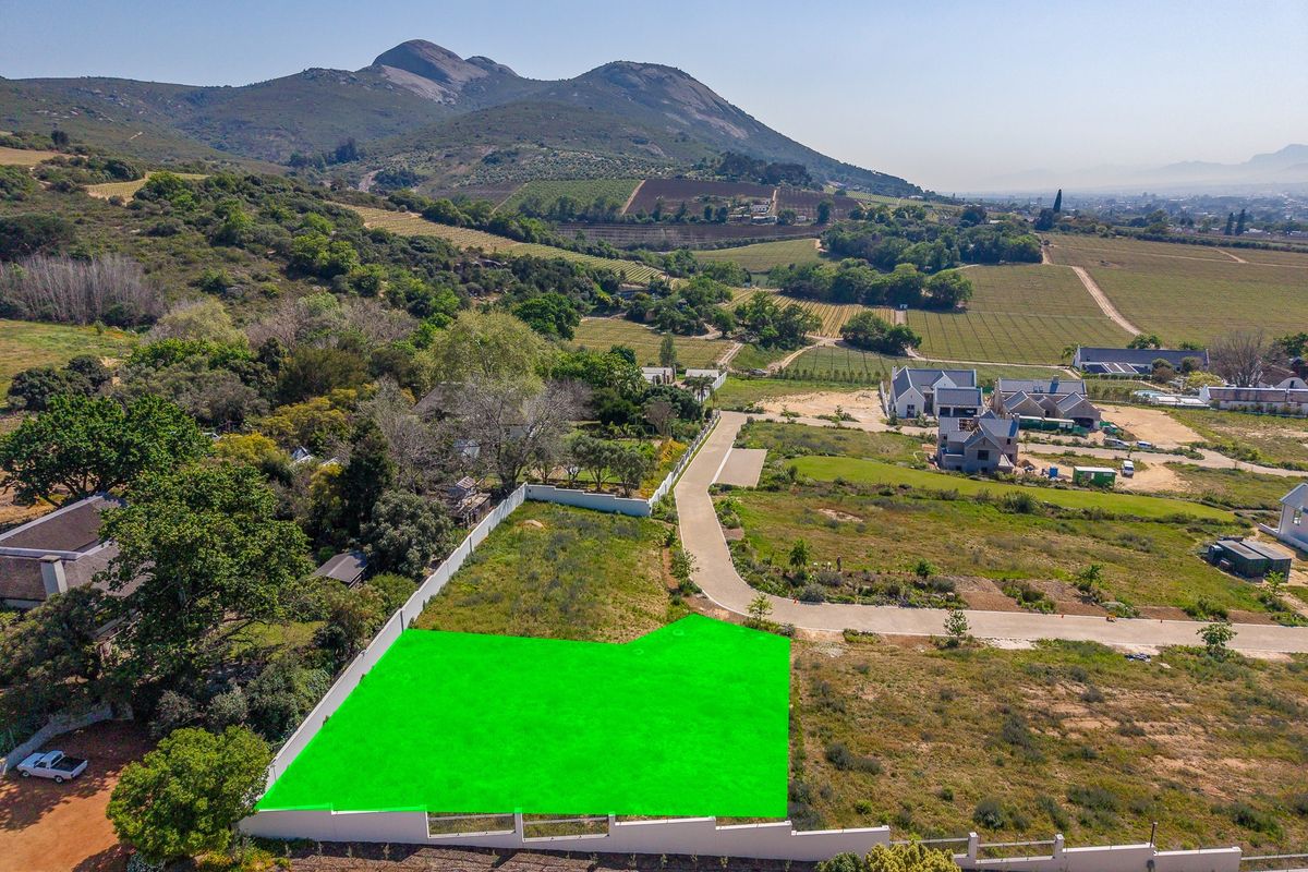 Aerial view of a vacant residential plot featuring a boundary wall and access to a paved road, set against a scenic mountain backdrop.