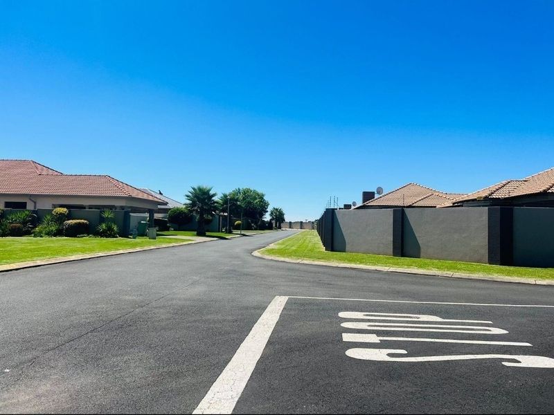 A clear street view of a residential estate featuring paved roads, well-maintained lawns, and concrete boundary walls under a clear blue sky.