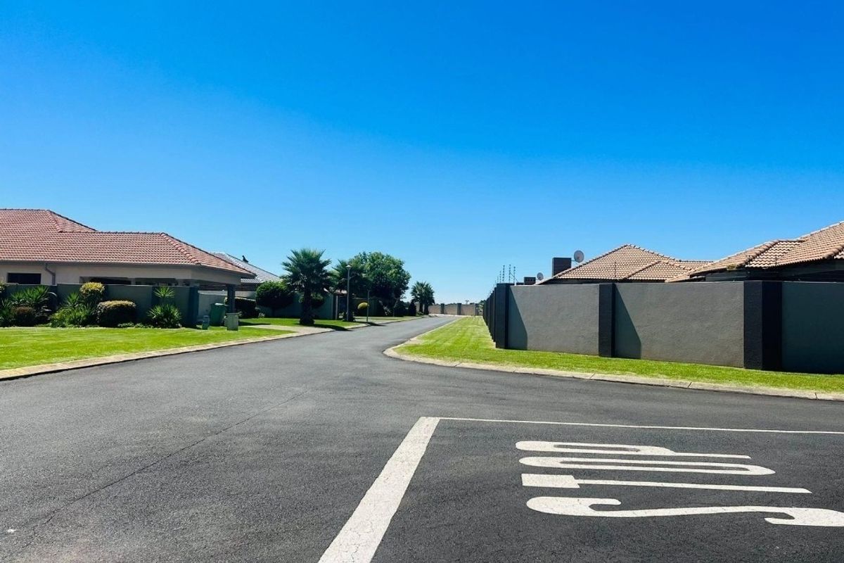 A clear street view of a residential estate featuring paved roads, well-maintained lawns, and concrete boundary walls under a clear blue sky.