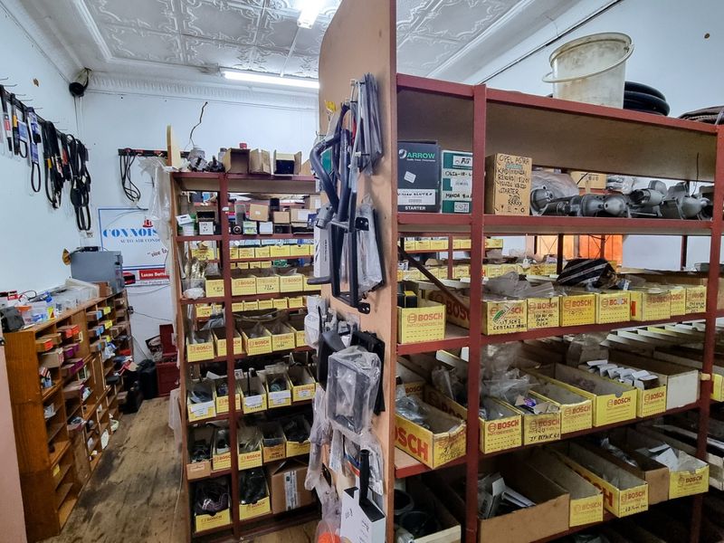 A commercial utility or storage space featuring industrial shelving units, pressed metal ceilings, and fluorescent lighting, currently utilized for automotive parts storage.