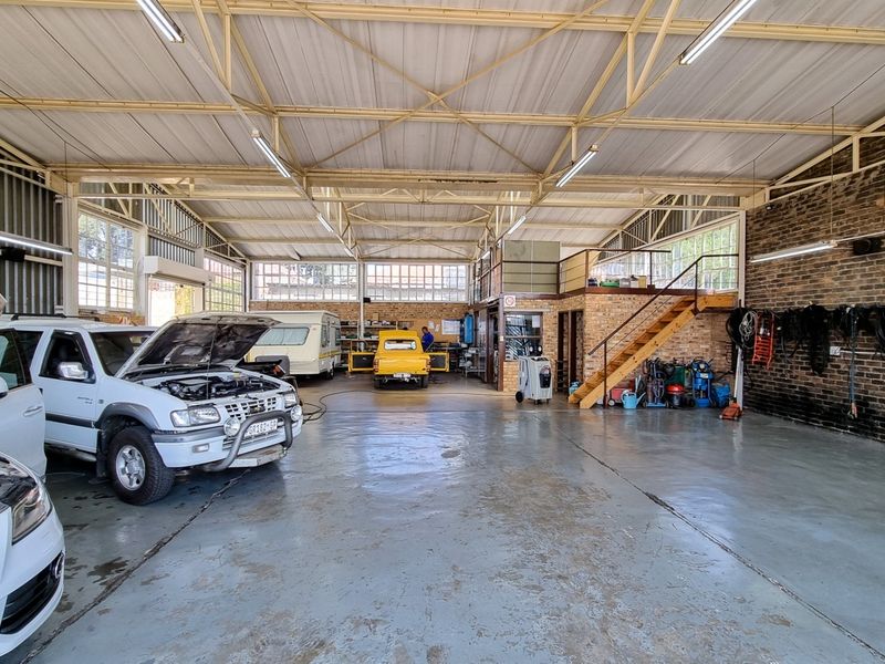 Spacious commercial garage workshop featuring exposed steel roof trusses, painted concrete flooring, exposed brick walls, and a mezzanine level with timber staircase access.