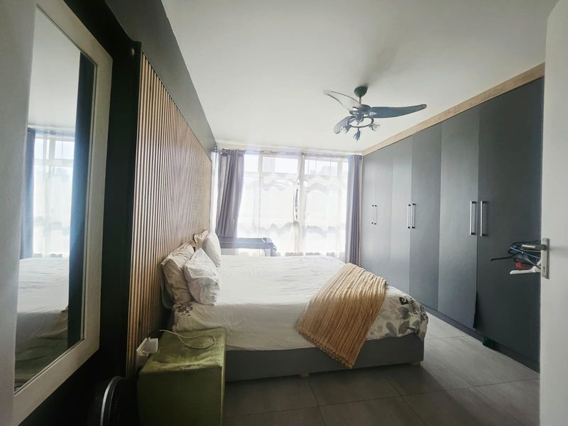 A modern bedroom featuring floor-to-ceiling built-in wardrobes, decorative slatted timber wall paneling, a ceiling fan, and neutral tiled flooring.