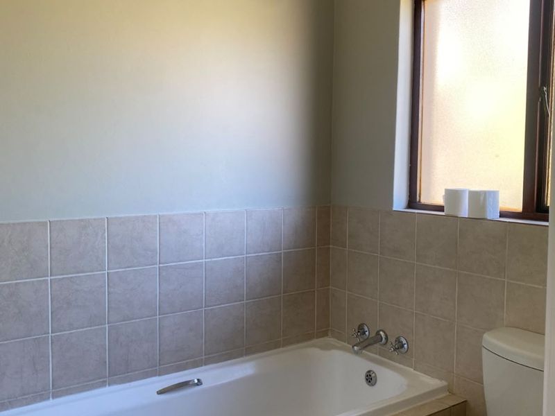 A functional bathroom featuring a built-in bathtub, pedestal basin, and toilet with neutral ceramic wall tiling and natural light from a frosted window.