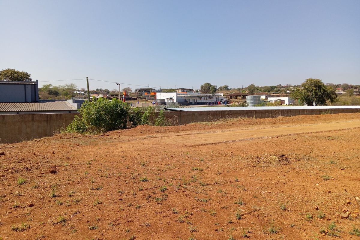 A view of the vacant commercial land showing the unpaved ground surface and a perimeter retaining wall with surrounding neighborhood buildings in the distance.