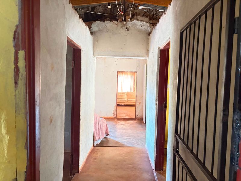 A narrow hallway featuring exposed ceiling rafters, concrete flooring, and a metal security gate, requiring significant renovation and finishing work.