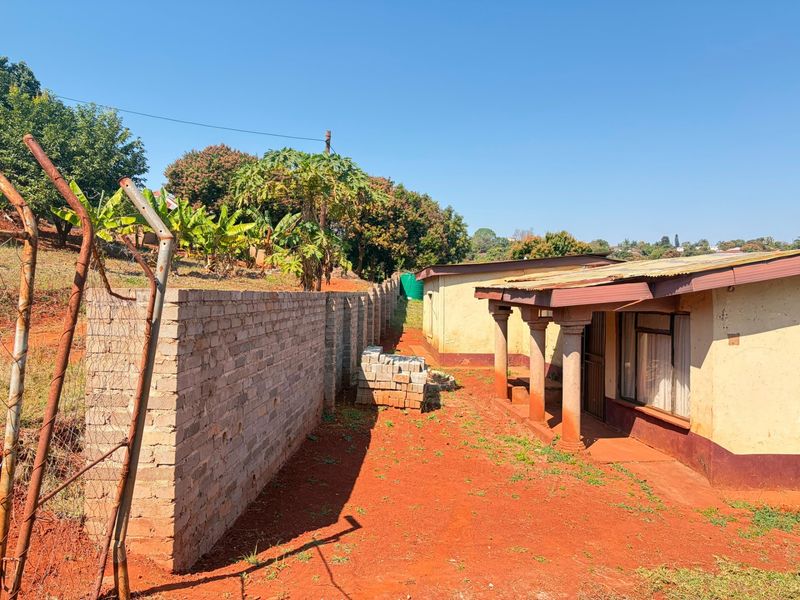 Rear exterior view of the property featuring a corrugated metal roof, concrete columns, and a brick boundary wall on an unpaved lot.