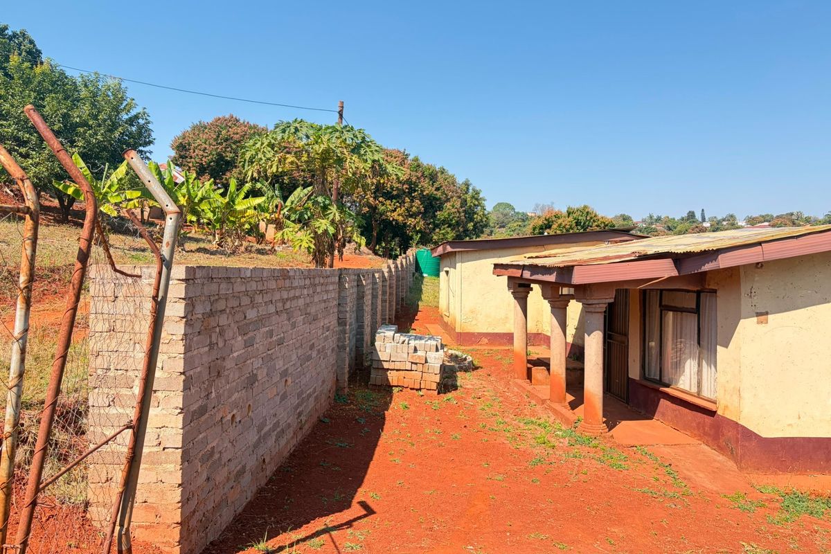 Rear exterior view of the property featuring a corrugated metal roof, concrete columns, and a brick boundary wall on an unpaved lot.