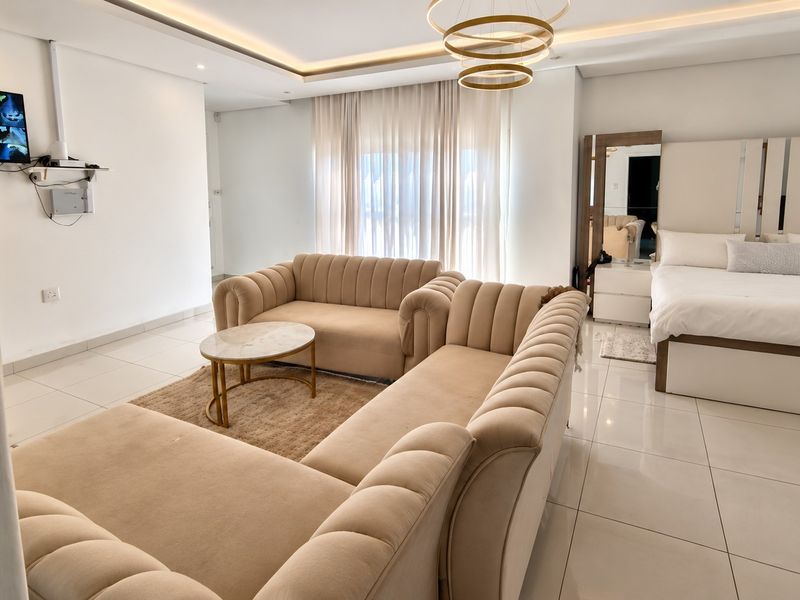 A modern open-plan living and sleeping area featuring neutral tones, tiled flooring, and recessed ceiling lighting.