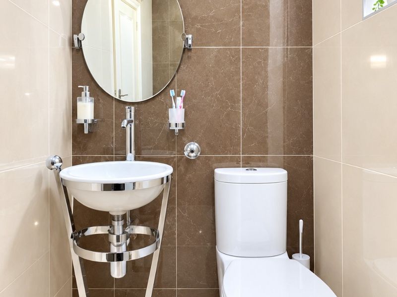 A modern, compact guest bathroom featuring high-gloss ceramic wall tiles, a contemporary wall-mounted basin with chrome stand, and a sleek dual-flush toilet.