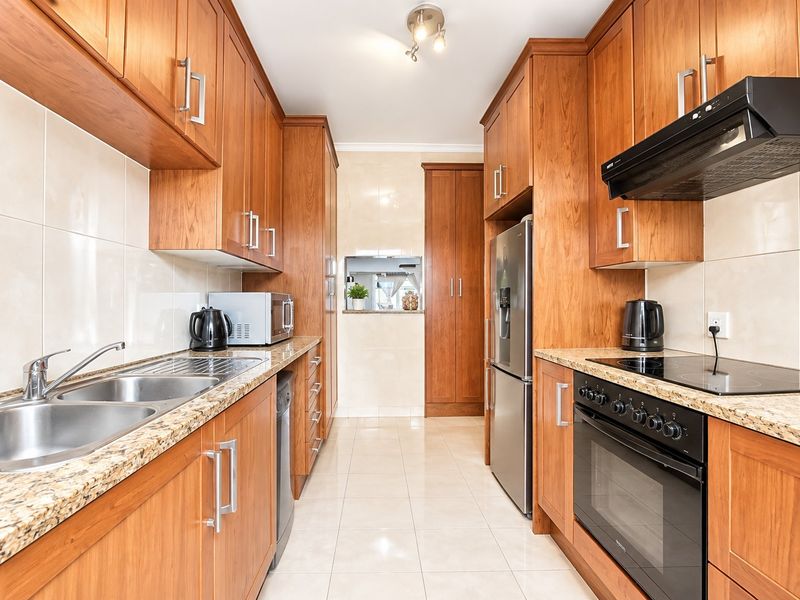 A well-maintained galley-style kitchen featuring warm wooden cabinetry, granite countertops, integrated appliances, and neutral tiled flooring.