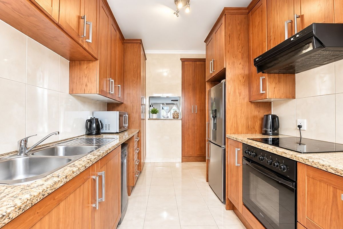 A well-maintained galley-style kitchen featuring warm wooden cabinetry, granite countertops, integrated appliances, and neutral tiled flooring.