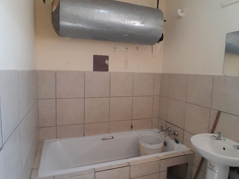 A dated bathroom featuring a bathtub, pedestal basin, and wall-mounted geyser, requiring cosmetic updates and repairs to wall tiling and plumbing fixtures.