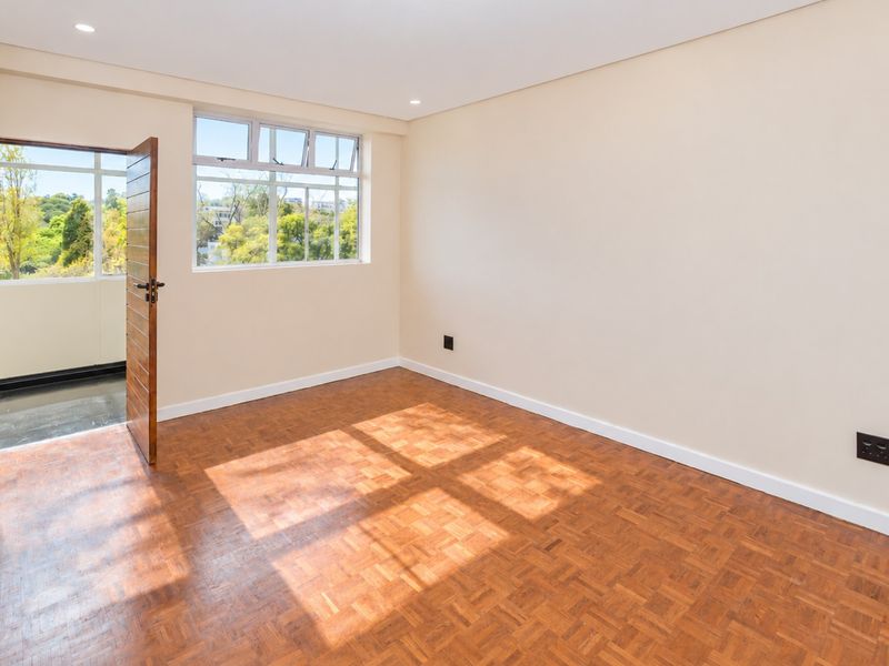 A bright, empty bedroom featuring classic parquet flooring, recessed lighting, and direct access to an outdoor balcony area.