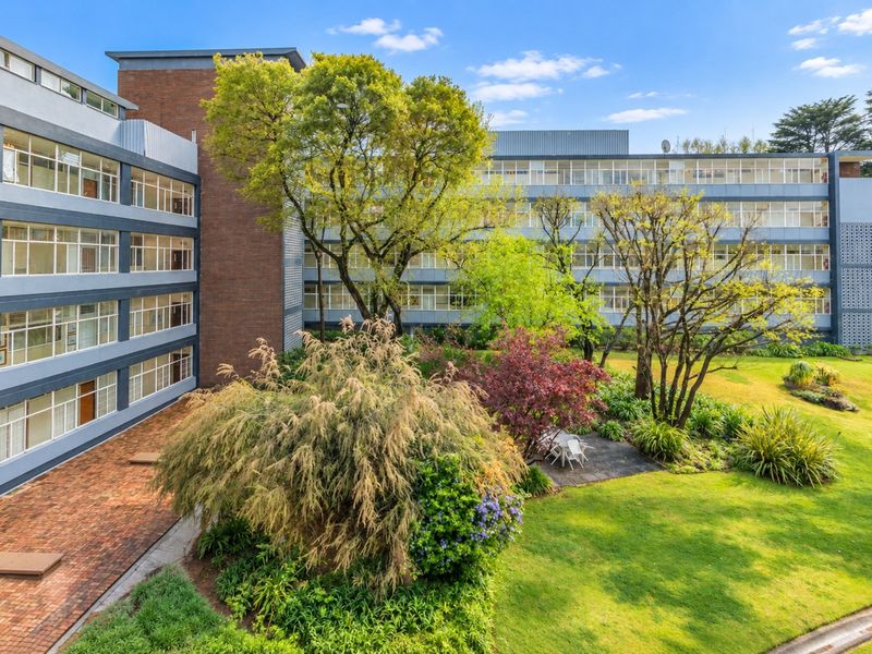 A view of the apartment complex exterior featuring a well-maintained garden, brick paved courtyard, and multi-story building facade with large windows.