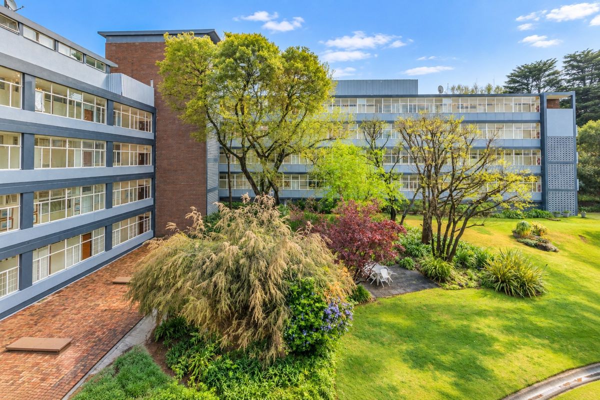 A view of the apartment complex exterior featuring a well-maintained garden, brick paved courtyard, and multi-story building facade with large windows.