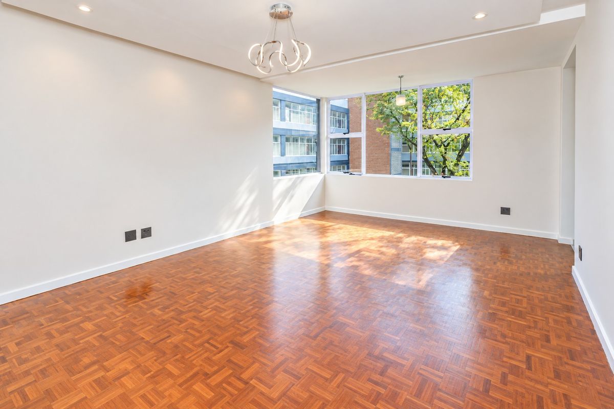 A bright and spacious living area featuring polished parquet flooring, modern lighting fixtures, and large corner windows providing ample natural light.