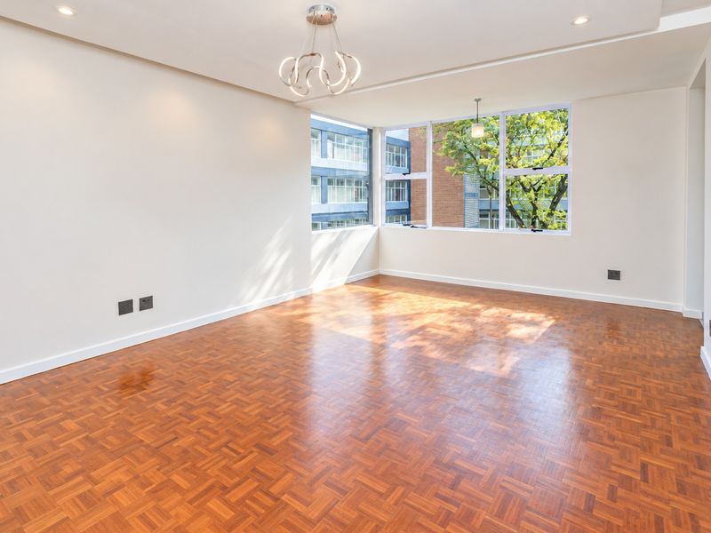 A bright and spacious living area featuring polished parquet flooring, modern lighting fixtures, and large corner windows providing ample natural light.