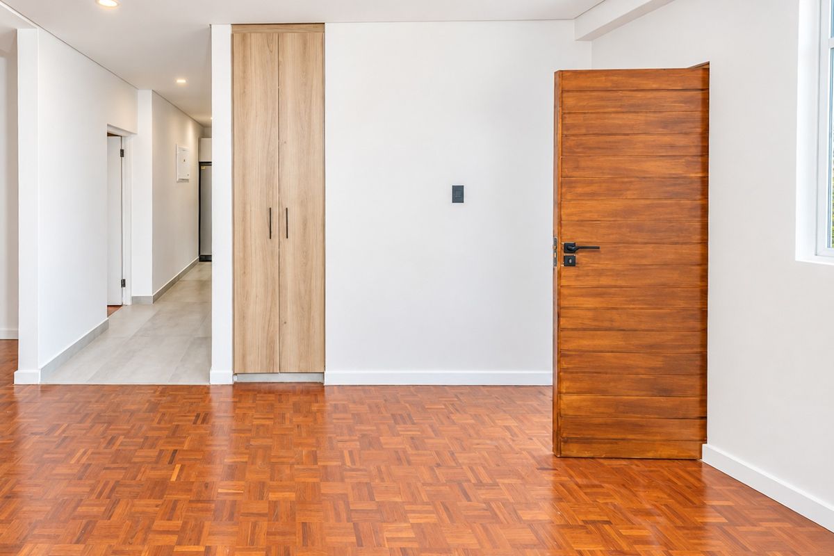 A bright, empty bedroom featuring polished parquet flooring, a modern built-in timber cupboard, and a solid wood door leading to a hallway.