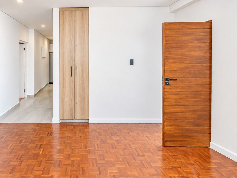 A bright, empty bedroom featuring polished parquet flooring, a modern built-in timber cupboard, and a solid wood door leading to a hallway.