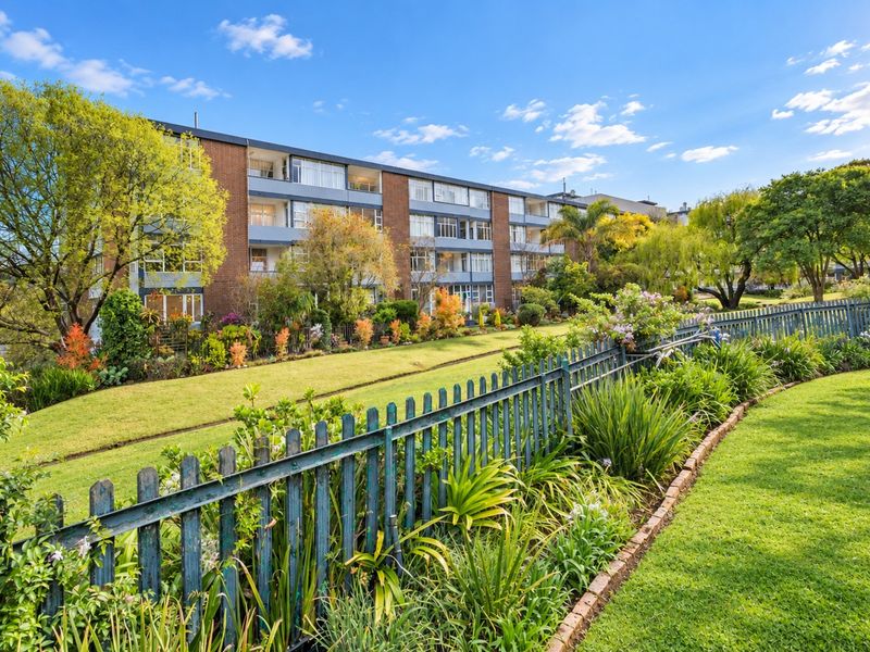 A view of the apartment building exterior featuring a well-maintained garden, lawn, and metal perimeter fencing.
