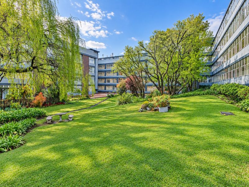 A well-maintained communal garden area featuring a lush green lawn, mature trees, and paved pathways surrounded by the apartment building complex.