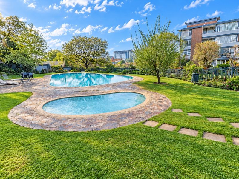 Well-maintained communal swimming pool area featuring paved surrounds, lush green lawns, and established garden landscaping within an apartment complex.
