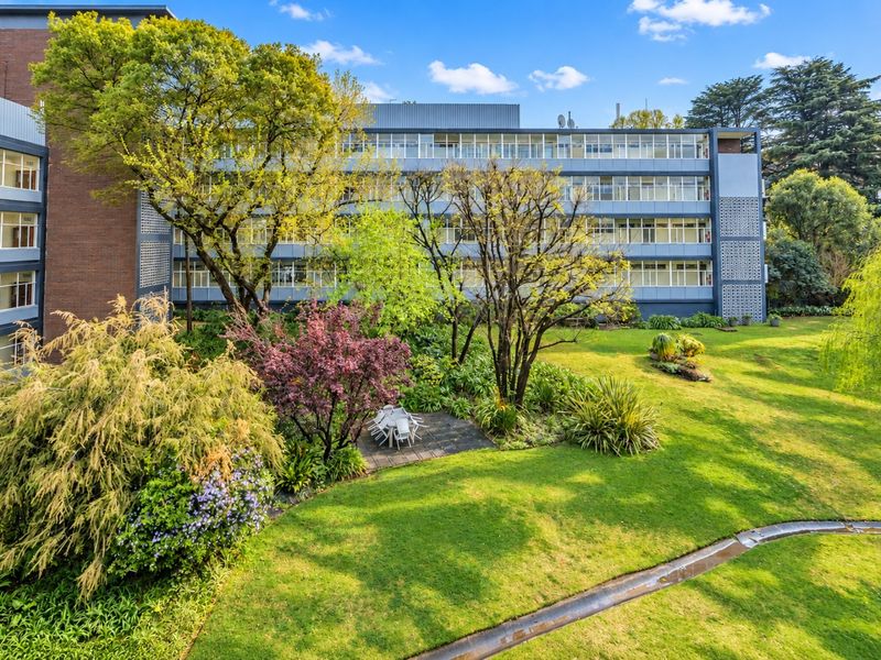 A view of the rear apartment building facade overlooking a well-maintained communal garden with mature trees and a paved seating area.